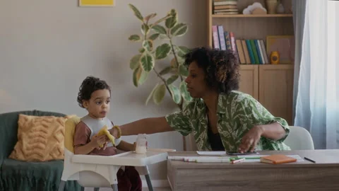 Mom Working at Home Table and Helping Little Son Eating Banana Stock Footage 334124242