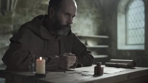 Monastic scribe praying with a rosary, gazing ahead, sitting at a wooden desk 스톡 동영상 337649363
