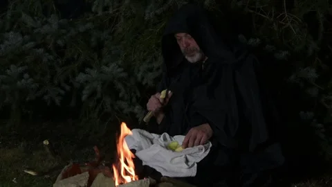 A monk in a black cassock is having dinner at night in the woods by the fire. Stock Footage 303783377