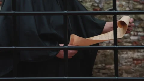 A monk in a black cassock unrolls a scroll and reads behind a grille against a s Video stock 264075383