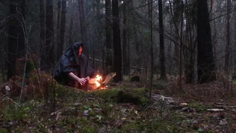 Monk in a black robe is warming himself by a fire in the dark autumn forest. Video stock 121935218