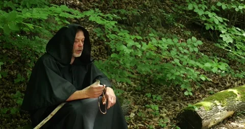 Monk in a dark cloak sits on a forest trail and holds a rosary during prayer. Stock Footage 336693628