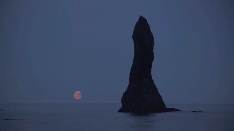 Moon rising low over calm ocean next to Reynisdrangar sea stack spire Iceland Stock Footage 111571240