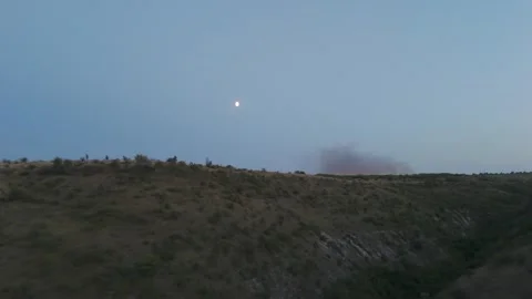 Moon rising over rolling hills with smoke plume in the distance, twilight sky Stock Footage 332725654