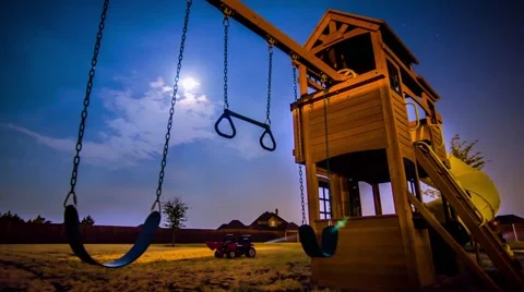 Moon setting over a fenced, backyard with a wooden tower swing set under a blue  Video stock 56801016