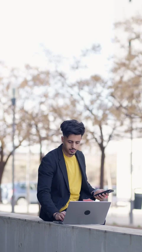 Moroccan man working remotely outdoors using laptop and smartphone. Vertical Stock Footage 343643793