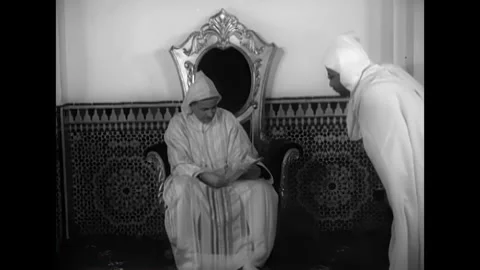 MOROCCO - 1951 - An advisor talks to Mohammed V of Morocco as he sits on his Stock Footage 340659911