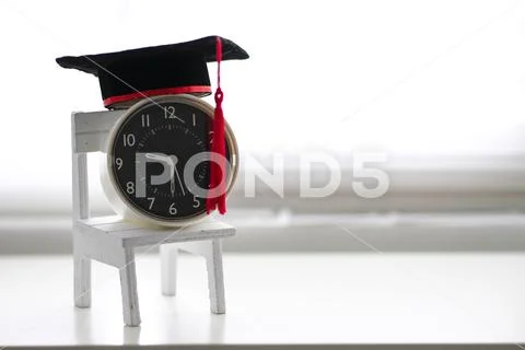 Mortarboard on an alarm clock and a toy chair, isolated in white background Stock Photos