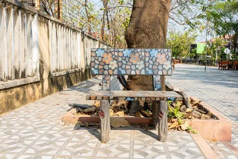 Mosaic tile park bench beneath a large tree in an outdoor public courtyard Stock Photos