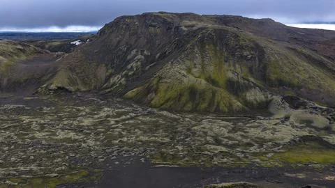 Moss-draped volcanic peak with deep erosion gullies overlooking dark lava fie 库存照片