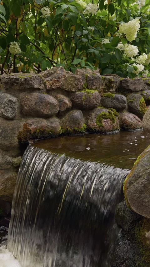 Mossy stone garden waterfall with flowing water, tranquil pond, and white Stock Footage 345455287