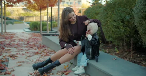 Mother adjusting toddler hat while sitting in autumn city courtyard Stock Footage 324577352