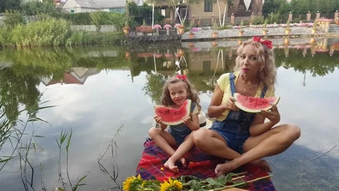 Mother and four-year-old blonde daughter eat watermelon on  the river Stock Footage 158932073