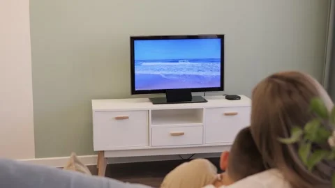 Mother and son sit on sofa, engrossed in TV. The television flickers, casting a Stock Footage 274790797