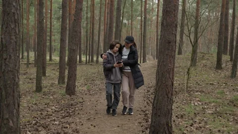 Mother and son using a smartphone together while walking arm in arm in a forest Stock Footage 316150898