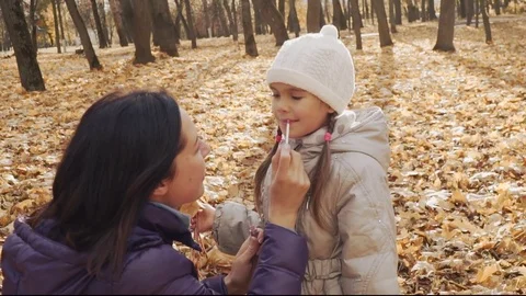 Mother is applying the gloss on the daughter's lips in the autumn park. Stock Footage 83504861