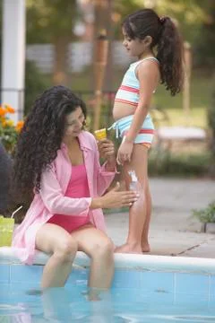 Mother applying lotion to daughter's leg at side of pool Stock Photos