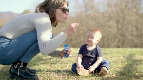 A mother crouches down to blow bubbles for her toddler, who sits on the grass in Stockbeeldmateriaal 278072864