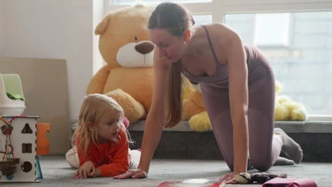 Mother does sport exercises while daughter plays at nursury room, workout Stock Footage 233194221