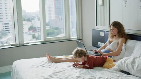 Mother is engaged with her laptop while her young child plays on the bed in a Stock Footage 296513009