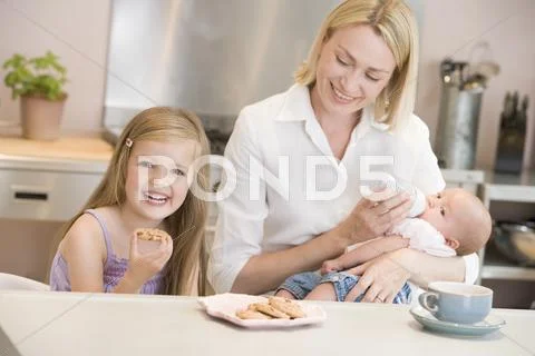 Mother feeding baby in kitchen with daughter eating cookies and smiling Stock Photos