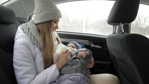 A mother feeds a one-year-old child in the back seat of a car with milk from a Stock Footage 162832983
