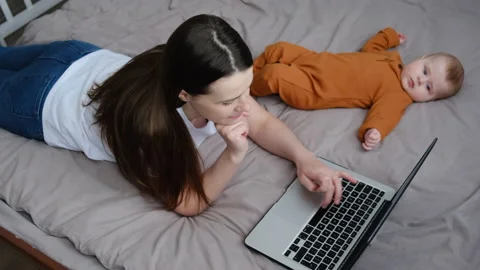 Mother freelancer working on computer lying on bed near baby infant at home Stock Footage 151084081
