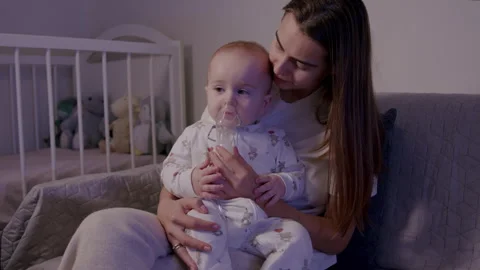 Mother gently holds infant using a nebulizer, showcasing a caring moment in a Stock Footage 323234225
