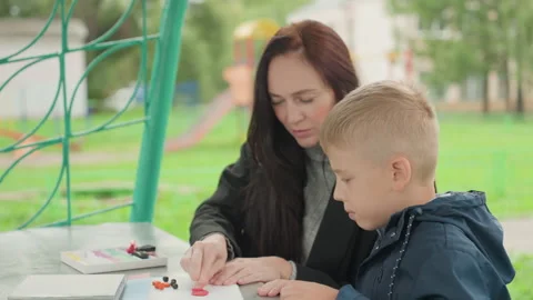 Mother guides craft. Child assembling beads outdoors. Park scene with mother and Stock Footage 335313359