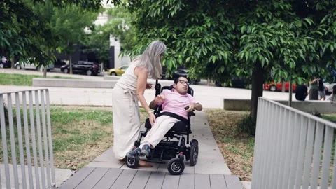 Mother helping her son with a physical disability in a wheelchair at the park Stock Footage 341256714