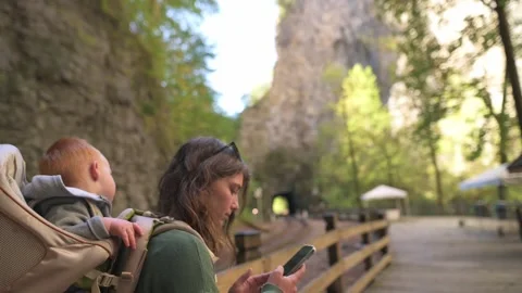 Mother with her child in backpack stops to check her phone while walking along 動画素材 295168491