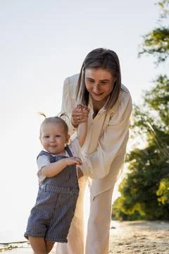 Mother holding baby's hands while teaching them to walk on a sunny beach Photos