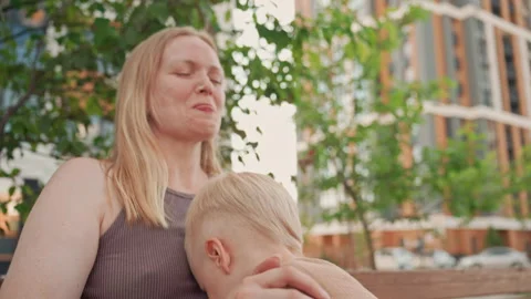 Mother holding young son on bench with greenery and city background featuring Stock Footage 345968139