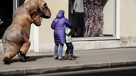 Mother holds her son while person in dinosaur costume running by at sidewalk Stock Footage 84928470