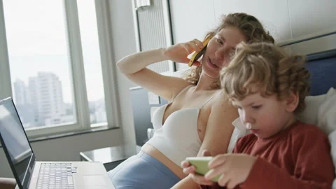 Mother multitasking on a call while her child plays on a mobile device in a Stock Footage 296512787