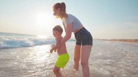 Mother plays with son at the beach during sunset while waves splash around them Stock Footage 338301034