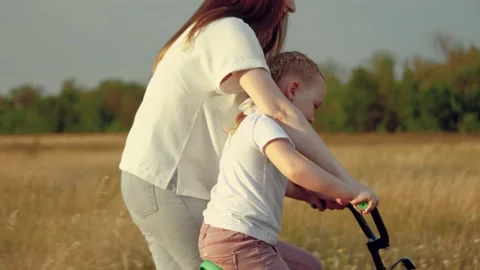 Mother pulls bicycle and little daughter pushes pedals in field at summer sunset Stock Footage 246598765