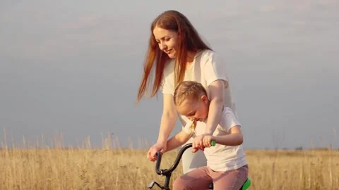 Mother pulls bicycle and little daughter pushes pedals in field at summer sunset Stock Footage 247488977