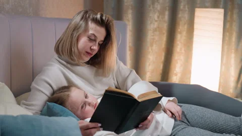 Mother reading book while daughter rests against her, cozy bedtime moment Stock Footage 325839884