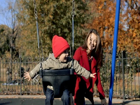 Mother in red coat pushing kid son on a swing. Slow motion Stock Footage