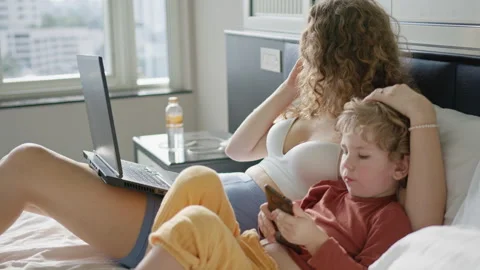 Mother sits on a bed talking on her phone while her child plays with a Stock Footage 296513446