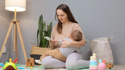 A mother sits comfortably on the floor while caring for her baby. She uses her Vídeo Stock 299939350