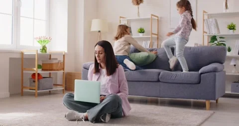Mother sits on floor working with laptop, kids play around making noise, home Stock Footage 334103164