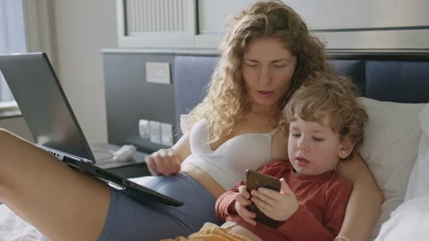 A mother is sitting on a bed with her son. She is working on her laptop while Stock Footage 296512316