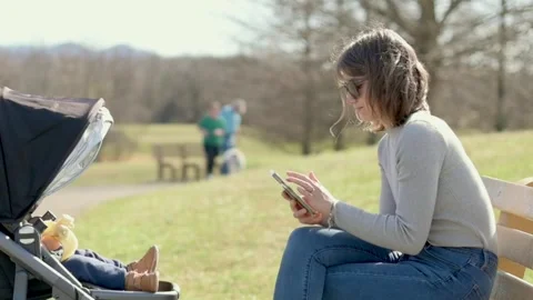 A mother sitting on a bench using her smartphone while her baby rests in a Vidéo 278017675