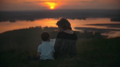 Mother with son sitting on the hill reading a book at summer sunset Stock Footage 89906512