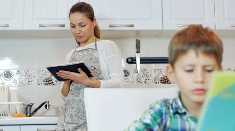 Mother standing in the kitchen with tablet computer. and son doing homework Stock Footage 44191987