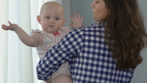 Mother standing by window dandling small child, kid looking at camera confused Stock Footage 80673013