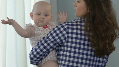 Mother standing by window dandling small child, kid looking at camera confused Stock Footage 80673161