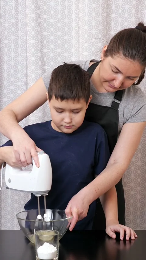 Mother teaching her smiling son how to cook, using a hand mixer to whisk egg Stock Footage 344798381